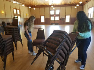 Picture of two students stacking chairs in a large event space with wooden floors