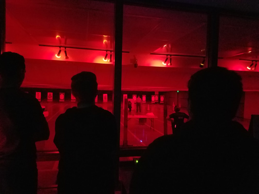 Picture of red lit room to simlulate the night time, students stand in a windowed room watching police cadets practice in a simulated crime scene