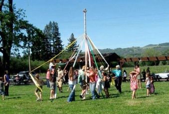 Picture of group of adults and children with ribbons in their hands dancing around a white May Pole. Each ribbon is attached to the pole so that as dancers go around the ribbons braid down the pole into an intricate braiding pattern. The dancers and May Pole are located on a grassy area with oak trees and hills in the distance