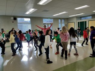 Picture of elementary school students in various poses of dance or stretch. The instructor is standing in the middle of the room with her arms stretched out
