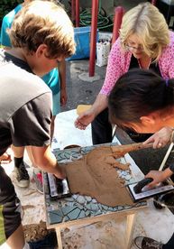 Picture of teacher and students working closely together grouting a mosaic table top. The two students are holding tools and spreading cement across the surface of the tile mosaic