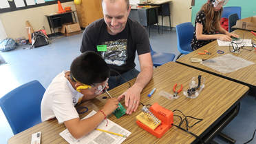 Picture of adult at table working with student wearing safety goggles. The table has various implements and tools on it
